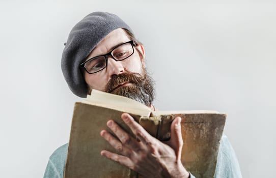 Portrait of serious bearded man in glasses and beret reading old book. Selective focus.