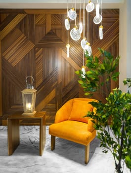 Orange armchair, illuminated wooden lantern on small wooden table, and contemporary tall glass chandelier, in a hall with decorated wood cladding wall, and white marble floor