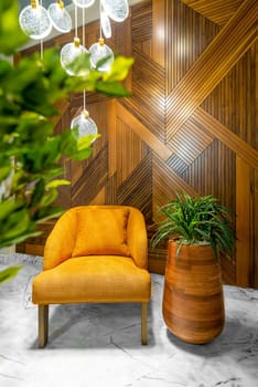 Modern orange armchair, tall wooden planter, and contemporary glass chandelier, in a hall with decorated wood cladding wall, and white marble floor, framed by green leaves