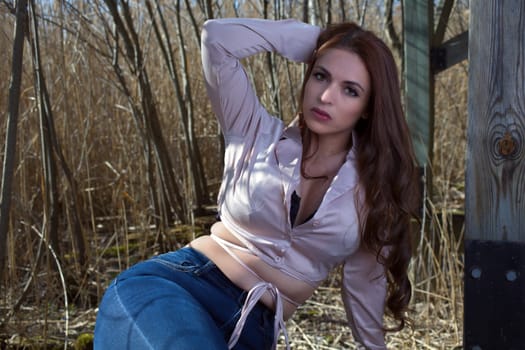 young woman brunette wearing pink shirt and jeans sitting in the forest holding her long brown hairs