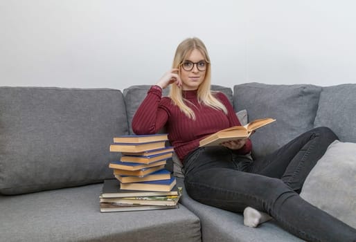 Young woman is sitting on the couch and reading a book leaning on a pile of books.