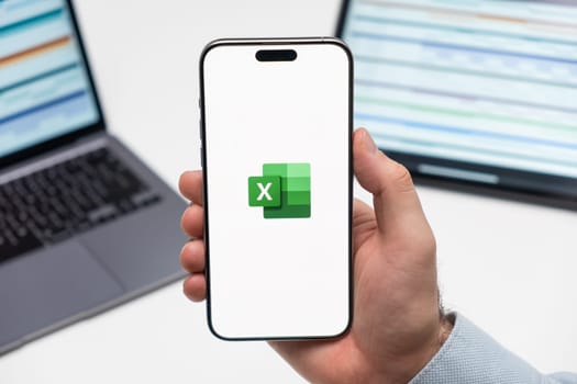 PRAGUE, CZECHIA - JULY, 2024: Excel logo on the screen of smartphone in mans hand and laptop on the background