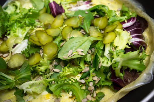 Hot salad over .scrambled egg on pan. Plate with several lettuces, gherkins and sunflowers seeds