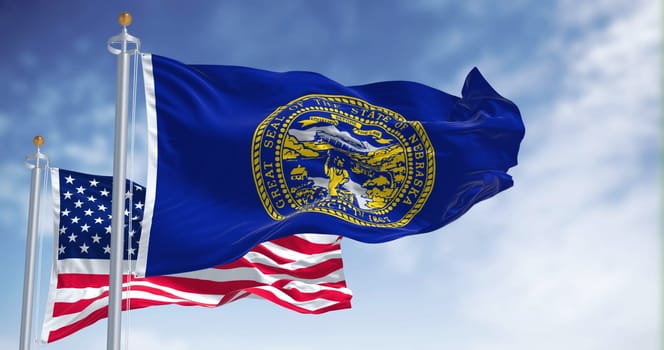 The Nebraska state flag waving along with the national flag of the United States of America. In the background there is a clear sky. Nebraska is a state in the Midwestern United States