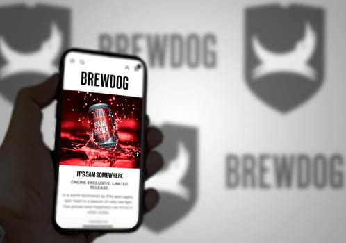 Ellon, Scotland, April 2022: The home page of the site Brewdog on the screen of a mobile phone. The mobile phone is held by a male hand. The company logo is blurry on the green background.