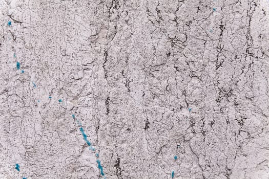 Brittle texture of a weathered ancient block of stone in an exterior load-bearing wall. Pale gray and light beige color. Black fissures, cracks and veins. Some blue paint stains. Flat background.