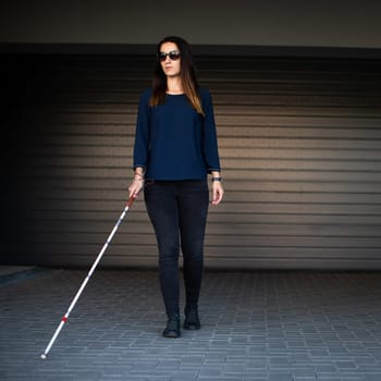 Young woman with impaired vision walking on city streets, using her white cane to navigate the urban space better and to get to her destination safely