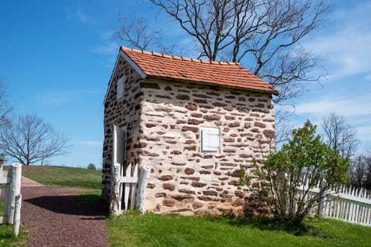 Colorful field stone smoke house with a rustic white wooden fence, blue sky and path with green trees and grass. Historic Pennsylvania architecture and early American construction.