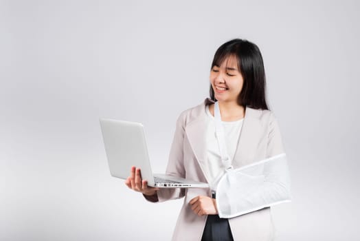 Asian business woman confident smiling broken arm after accident and wear arm splint for treatment but still have to work she hold laptop computer studio isolated on white background,copy space