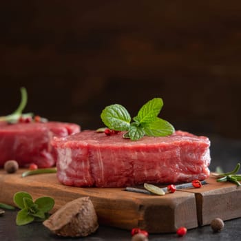 Raw beef fillet steaks on wooden board on dark table close up with copy space. Portioned filet mignons with spices and herbs. Concept of summer food, meat eaters.