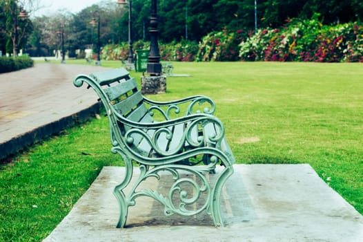 Side view of a garden chair, outdoors next to a footpath road and green grass field of a public park at sunny day during sunset. Nature background