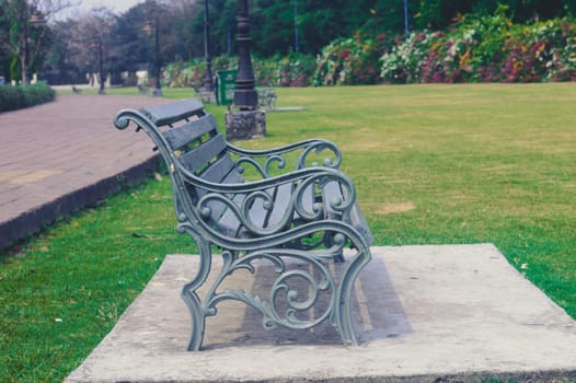 Side view of a garden chair, outdoors next to a footpath road and green grass field of a public park at sunny day during sunset. Nature background