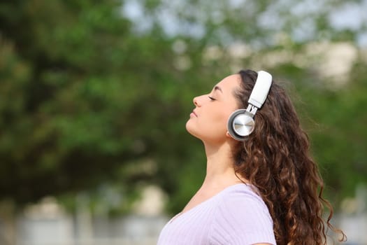 Side view portrait of a relaxed woman wearing headphones listening audiobook meditating in a park