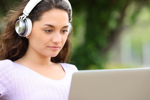 Concentrated woman with wireless headphones watching media on laptop in a park