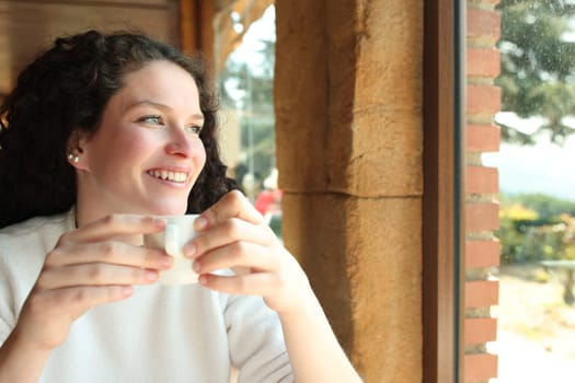 Happy woman drinking coffee sitting in a restaurant looking away through a window