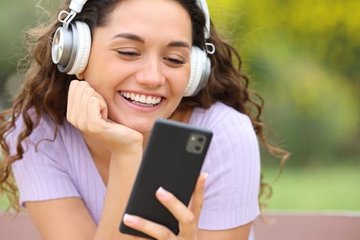 Front vie wportrait of a happy woman listening and watching videos on phone sitting in a bench