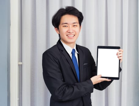 Work from home, Asian young businessman wear suit video conference call or facetime he smiling and showing blcnk screen of smart digital tablet computer at home office, looking to camera