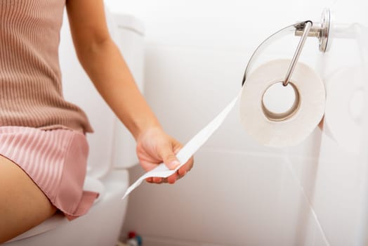 Closeup hand pulling toilet paper roll in holder for wipe, woman sitting on toilet she taking and tearing white tissue on wall to towel clean in bathroom, Healthcare concept
