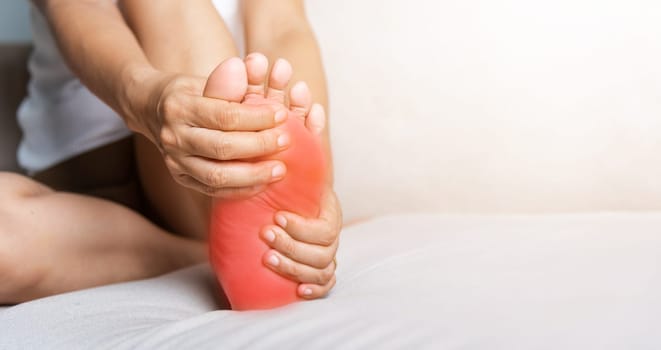 Foot pain, Asian woman sitting on sofa feeling pain in her foot at home, female suffering from feet ache use hand massage relax muscle from soles in home interior, Healthcare and podiatry medical