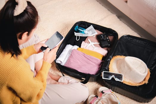 Preparing for travel in new normal. Woman chatting surfing internet use smartphone application during packing suitcase baggage, Female texting message on mobile phone with friend for ready
