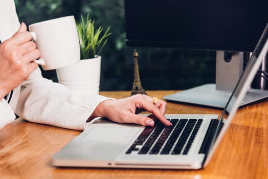 Beautiful business woman lifestyle working using laptop computer and holding coffee cup on hand while enjoying in modern desk office near window, work from home concept