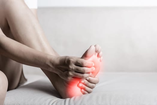 Foot pain, Asian woman sitting on sofa feeling pain in her foot at home, female suffering from feet ache use hand massage relax muscle from soles in home interior, Healthcare and podiatry medical