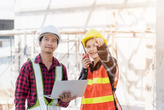 Asian engineer foreman worker man and woman working at building construction site use laptop and talking with radio, engineering hold computer and radio discuss and control worker employee to building