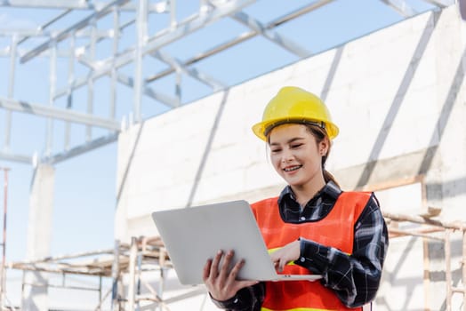 Asian engineer architect worker woman holding laptop inspect and oversee infrastructure progress at construction site, engineering use computer to operate project and control worker to building