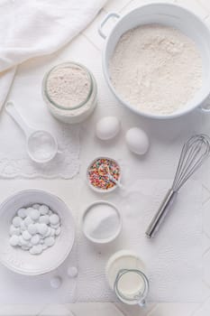 Baking background with flour, eggs, sugar sprinkles, kitchen tools, utensils in white