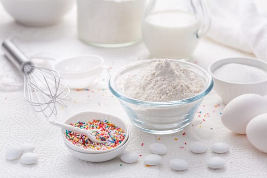 Baking background with flour, eggs, sugar sprinkles, kitchen tools, utensils in white