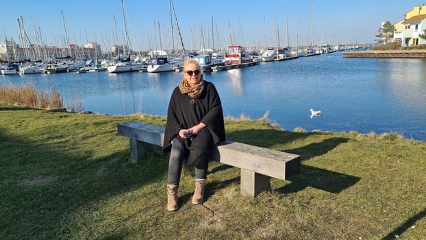 Portrait of a blonde woman wearing sunglasses sitting on a bench in the marina with boats in the background