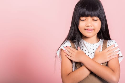 Asian little kid 10 years old doing grateful gesture and closed eyes at studio shot isolated on pink background, Happy child girl smile putting crossed palm on her chest, being thankful, Gratitude day
