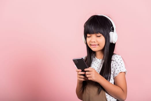 Asian kid 10 years enjoying listening music from mobile phone wear wireless headset closed eyes at studio shot isolated on pink background, Happy child girl use smartphone listen music with headphones