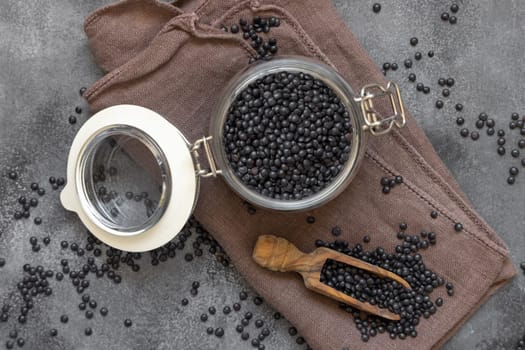 Glass jar of dry black lentils beans with a wooden scoop on grey table top view. Healthy eating, protein source for vegan and vegetarian diet. Traditional Asian and North African cuisines ingredient