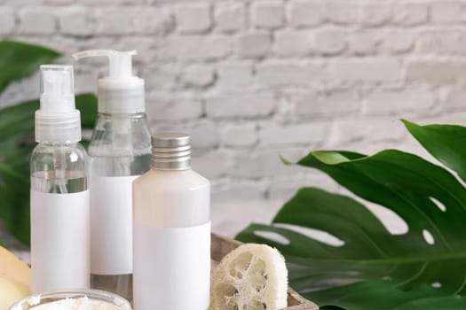 Cosmetic bottles and accessories near green monstera leaves close up against white brick wall. Brand packaging mockup. Skincare beauty products. Natural homemade cosmetic ..