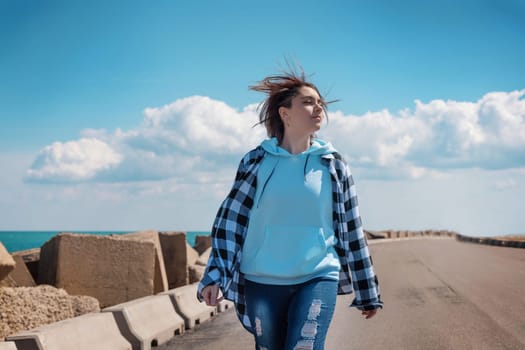 Short haired teenager in light blue hoodie and plaid shirt. Teen girl walking outdoor on the road and looking down, sunny day and blue sky. Clothing mockup, freedom adolescence concept