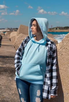 Teenager in light blue hoodie and plaid shirt looking aside. Teen girl stays outdoor near the sea, concrete blocks and blue sky, windy day. Clothing mockup, copy space