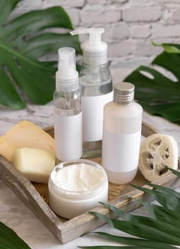 Cosmetic bottles and skin care accessories near green monstera leaves close up against white brick wall. Brand packaging mockup. Organic beauty products. Natural homemade cosmetic ..