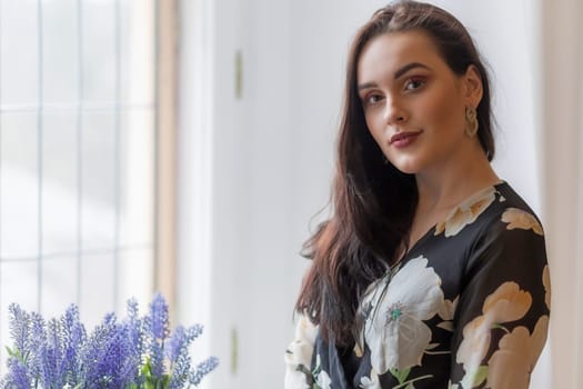 Attractive dark haired girl is posing next to purple flowers in windows. Horizontally.