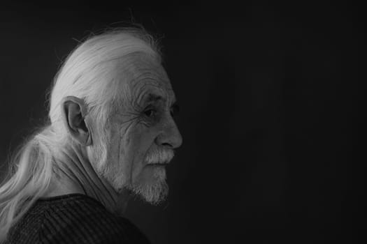 Low key studio black and white portrait of beautiful gray hair old man looking over his shoulder at the camera. Horizontally.