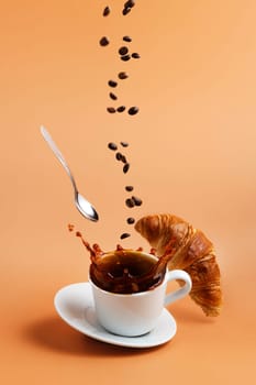 White coffee cup, plate and metal tea spoon falling on beige table background and coffee splashes because of hit, coffee beans levitate, fly and fall to cup, crispy fresh croissant. Breakfast concept