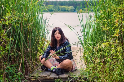 A black-haired young girl in a black jacket sits on a wooden bridge on the lake shore