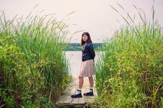 a brunette girl in a dress and a black jacket stands on the river bank on a wooden bridge among the reeds