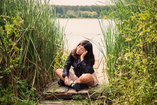 a brunette girl in a black jacket sits on a wooden bridge on the bank of a pond, propping her head up with her hand