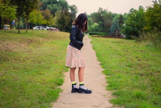 young woman with long dark hair looks at her shoes while standing on a path in the park