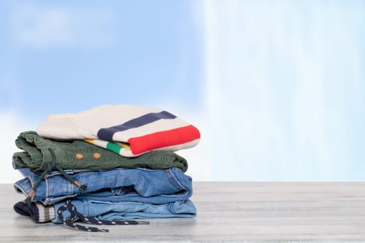 Stack of jeans. Closeup of a pile of colorful denim pants and one colorful sweater on a bright table against abstract blurred light blue curtain background.. Template for your product display montage.