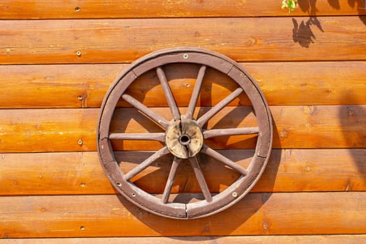 An old wooden carriage wheel with metal fittings hangs on the wall of a wooden house for decoration. Wooden spoke wheel. Outdoor natural light. Macro.