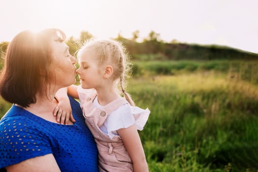 Grandmother holding granddaughter on her arms and kissing nose. Happy childhood, family love concept. Support and togetherness. Two people outdoor with sunlight background. High quality photo