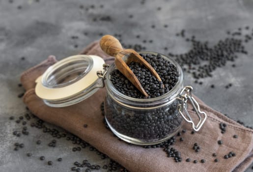 Glass jar of dry black lentils beans with a wooden scoop on grey table close up. Healthy eating, protein source for vegan and vegetarian diet. Traditional Asian and North African cuisines ingredient