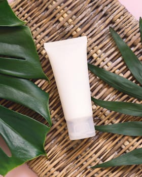 White Plastic tube lying between tropical leaves on wicker table top view. Brand packaging mockup. Natural herbal cosmetics concept - skincare beauty product, cream or lotion..